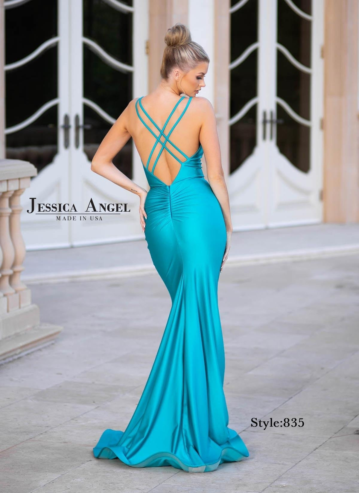 A woman stands outdoors near arched double doors, wearing the Jessica Angel 835 Cut-Out Long Mermaid Prom Dress in turquoise with crisscross straps and an open back.